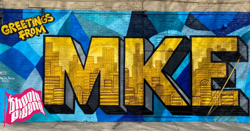 A mural in blue and yellow by Fred the Artist, also known as Fred Kaems, reads “Greetings from MKE!” The mural is on a wall of a business called Soda Sign. MKE is an abbreviation for Milwaukee, Wisconsin, USA. Inside the large gold block sans-serif letters “MKE” are glimpses of Milwaukee’s skyline.