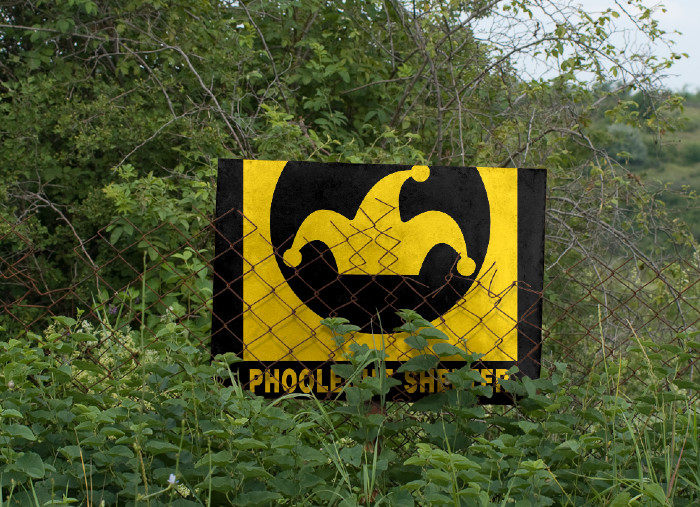 A PhooleOut Shelter sign has blown into an overgrown wasteland and is draped over a rusting, falling-down chain-link fence.